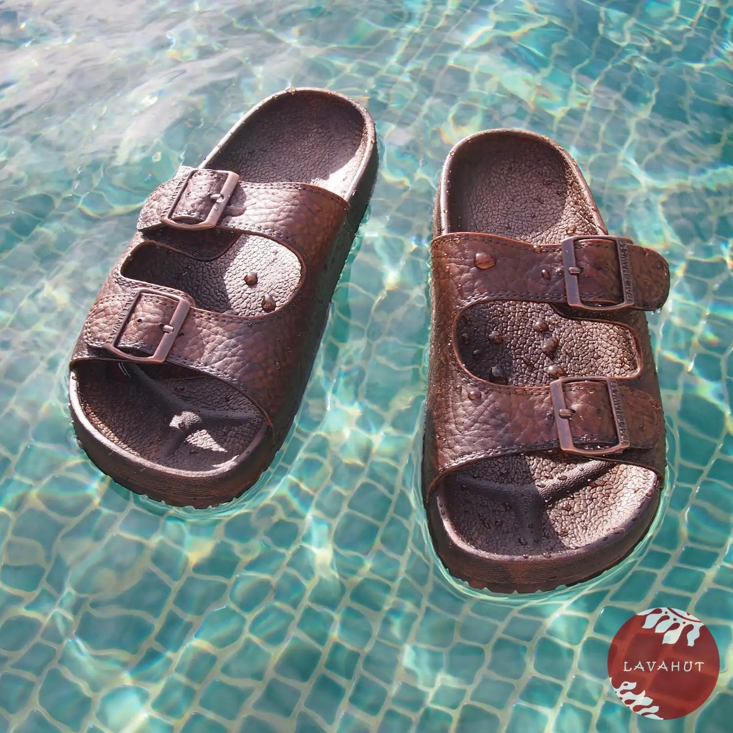 Brown leather-like sandals with two adjustable straps and metal buckles, floating in clear pool water.