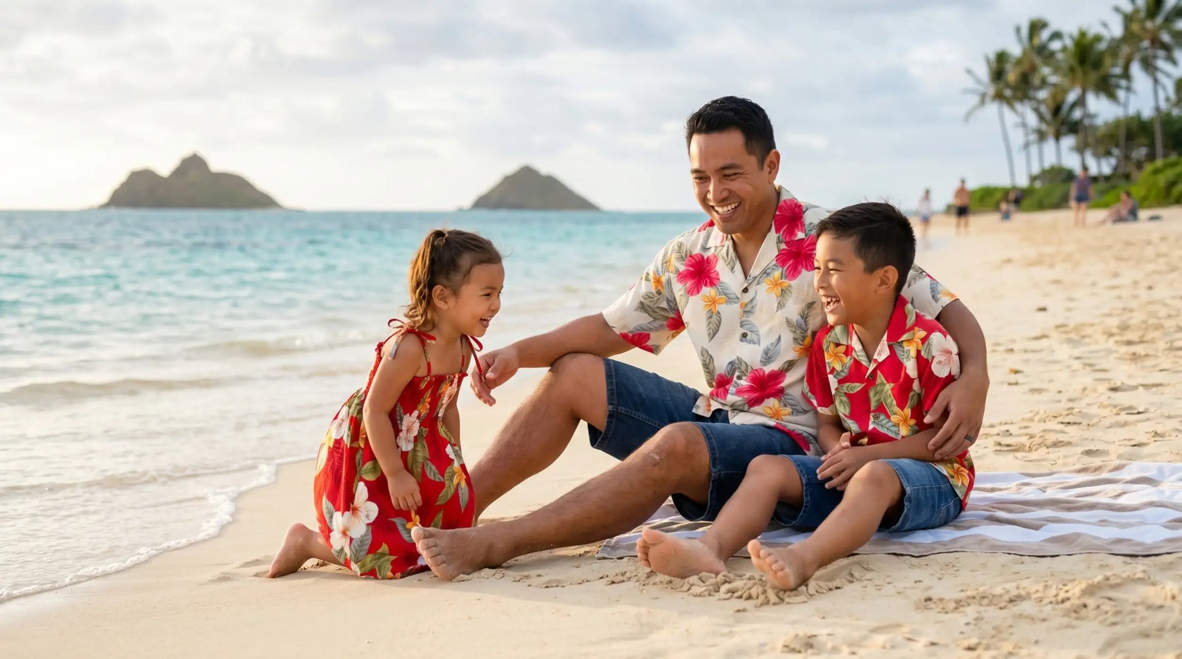 A man and two children wearing matching floral hawaiian shirts sit on a beach towel, with the father in a white shirt featuring red and pink flowers paired with blue shorts.