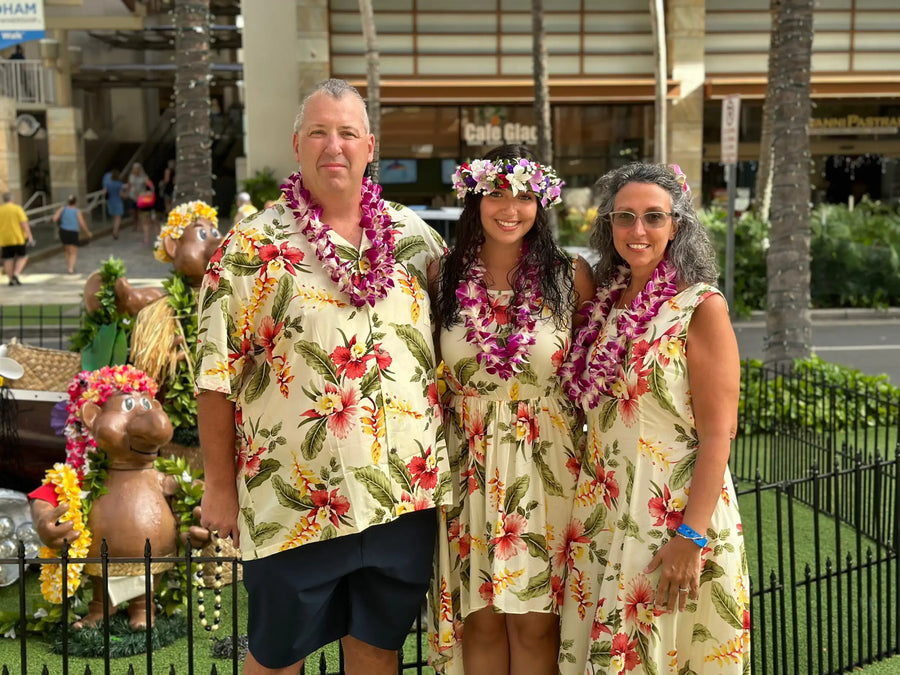 The three people are wearing matching cream-colored hawaiian shirts and dresses with vibrant red, pink, yellow, and green tropical floral patterns.