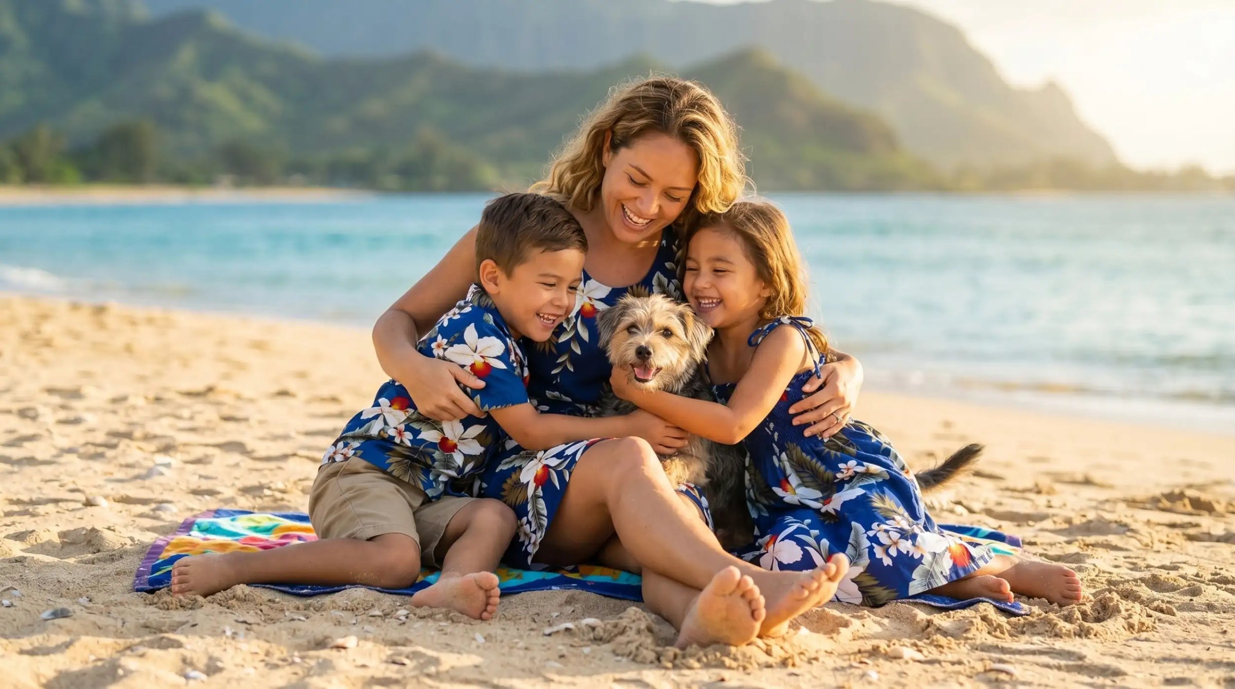 A woman and two children in matching blue floral hawaiian shirts sit on a beach towel with their small dog, all smiling together.